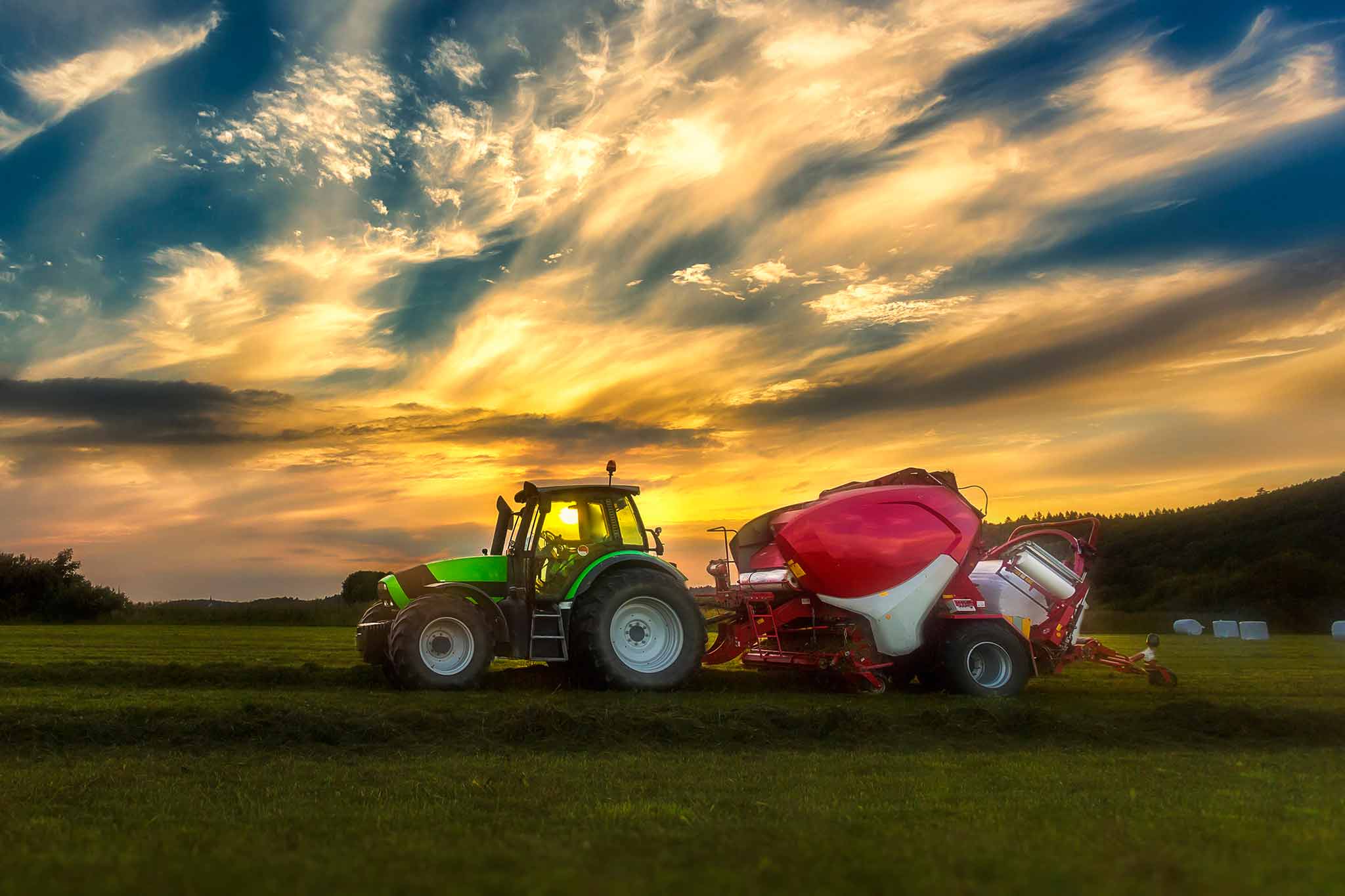 Vor einem Wolkenhimmel ist auf einer Wiese ein Traktor zu sehen, der eine Rundballenpresse angehängt hat. 