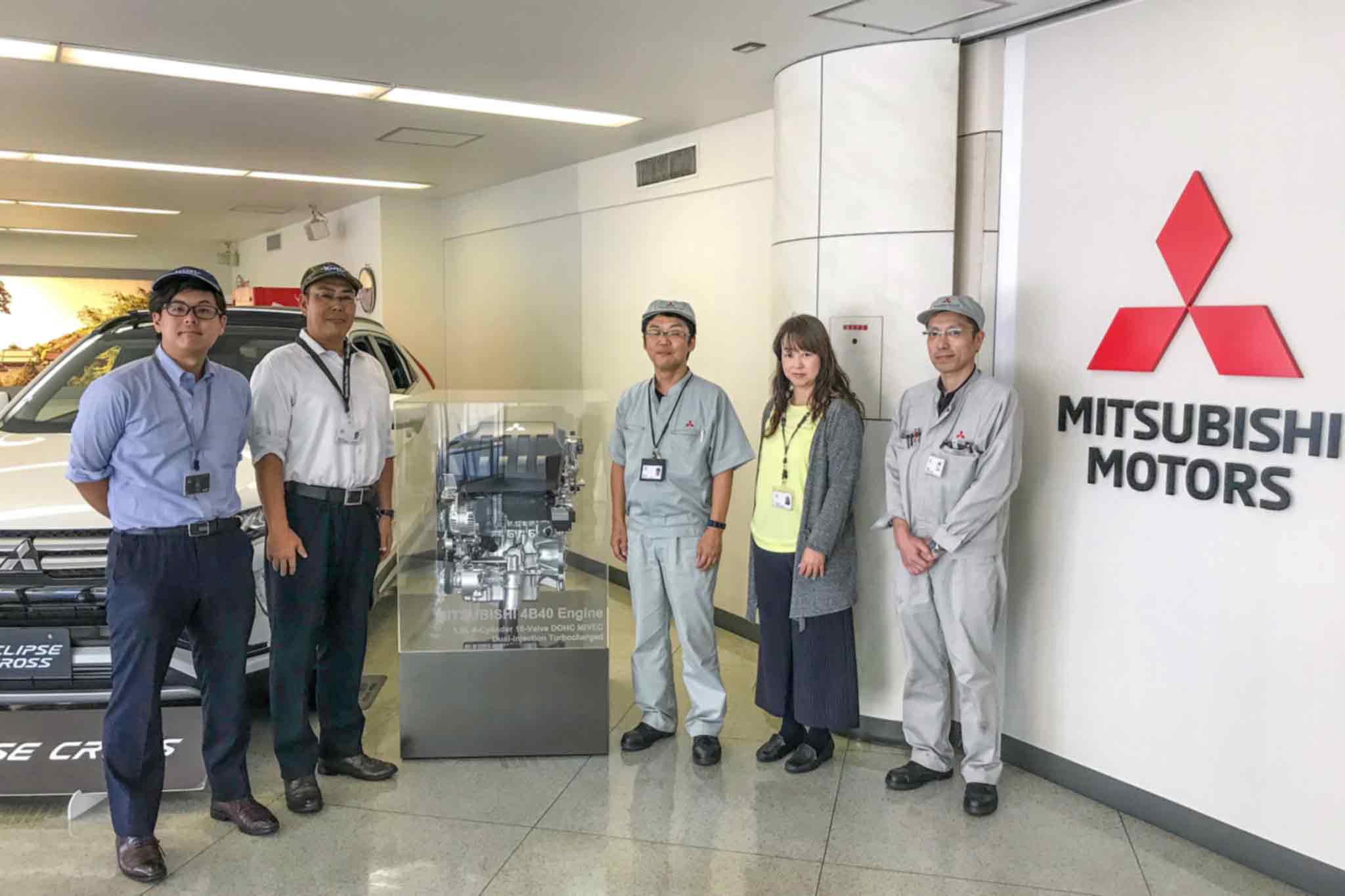Rechts das Mitsubishi Motors Logo. Links davon sind 5 Personen um einen Motor platziert. Im Hintergrund steht ein Auto.