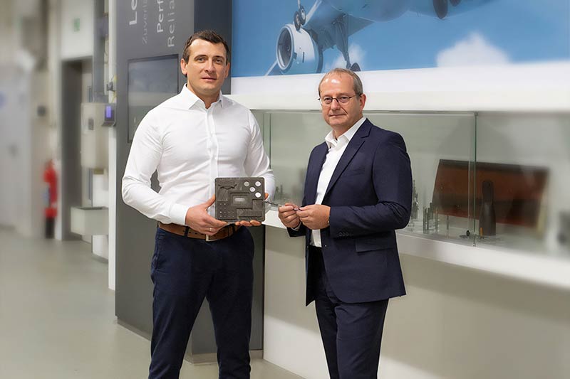 Two men stand in a presentation room. In their hands they hold a component made of CFRP and a drill.