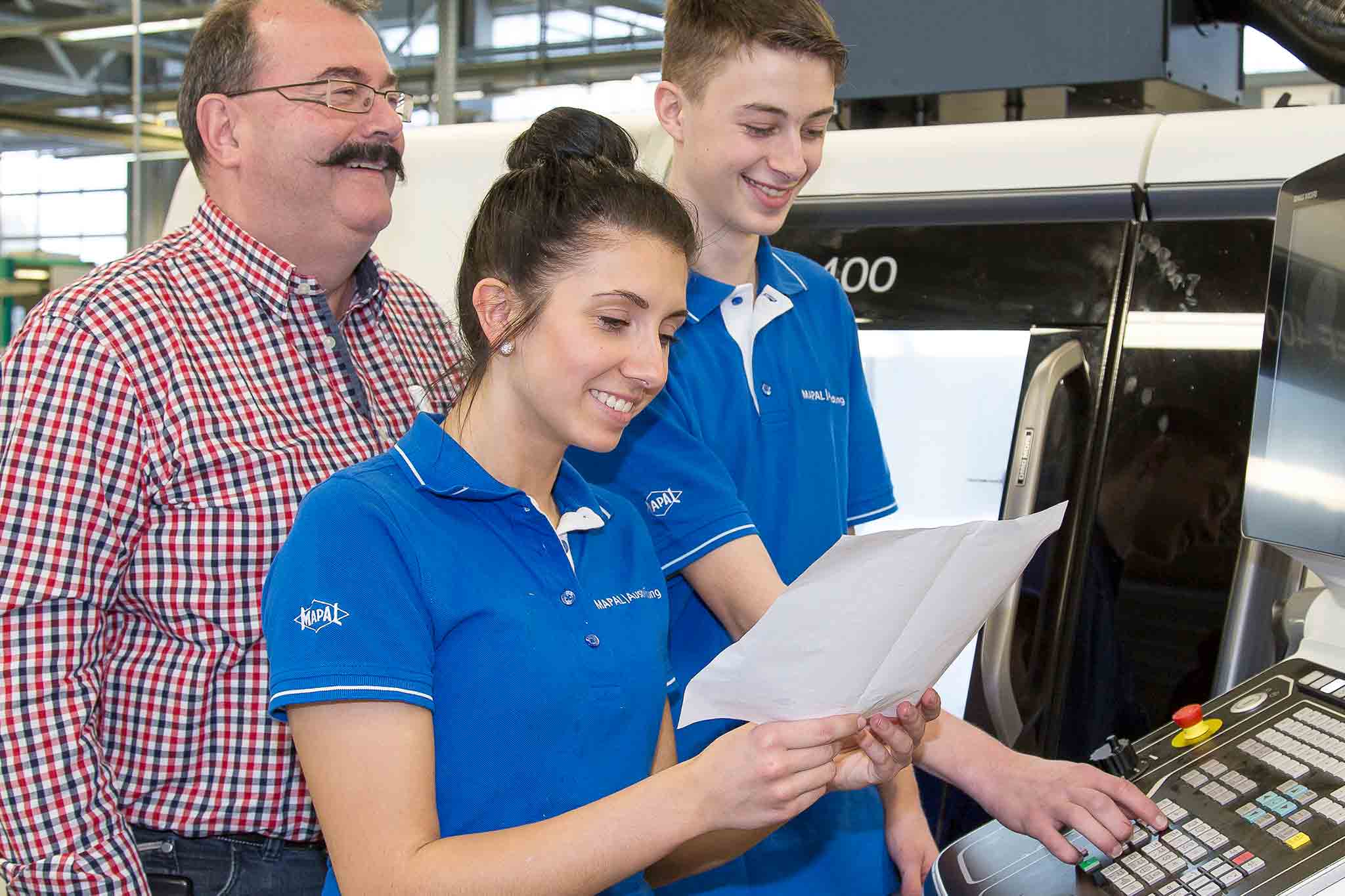 Zwei Auszubildende sind an der Maschine zu sehen. Ein technischer Ausbilder erläutert einen Sachverhalt.