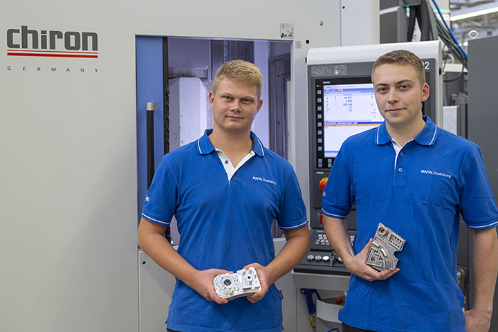 Julian Kraus and Lukas Weiss (from left) in front of a Chiron machine. Both of them are holding a part in their hands.