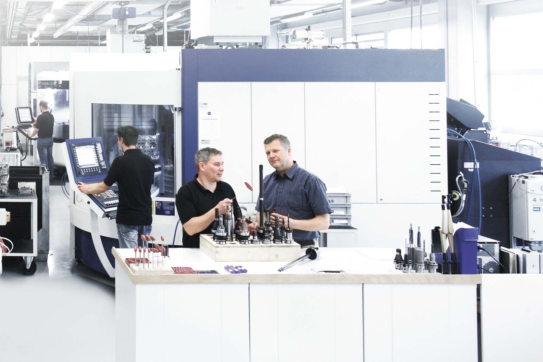 Two employees discuss the properties of various tools while other employees make adjustments to machines in the background.